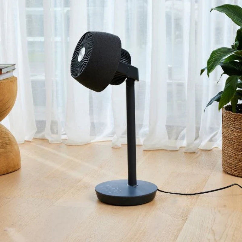 Black stand fan on a wooden floor with a plant and curtain in the background