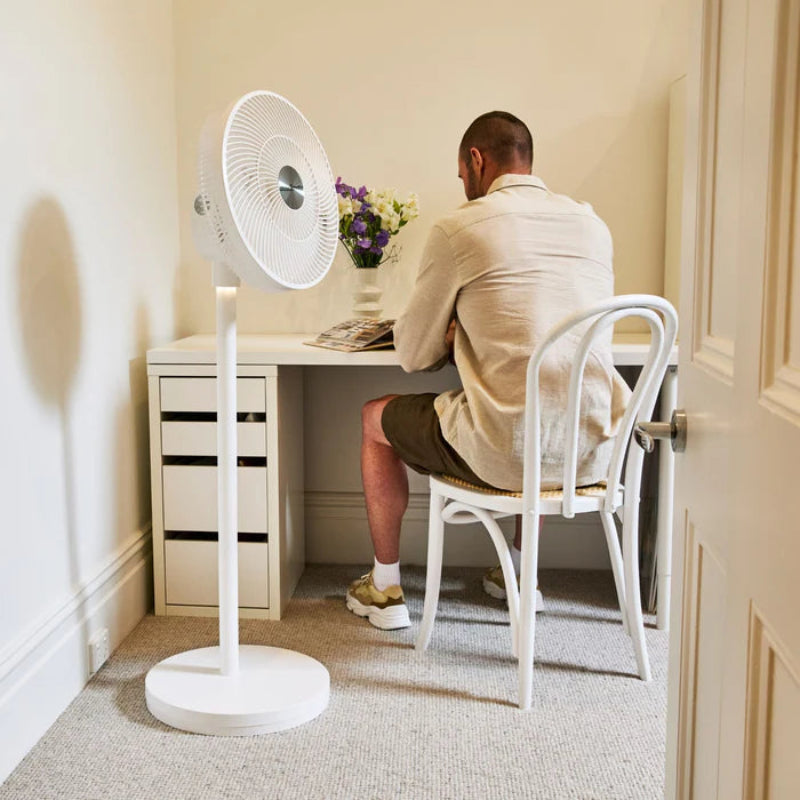 Person sitting at a desk with a white floor fan in a home office setting.