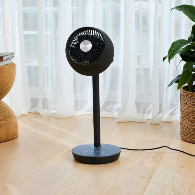 Black stand fan on a wooden floor with a plant and curtain in the background