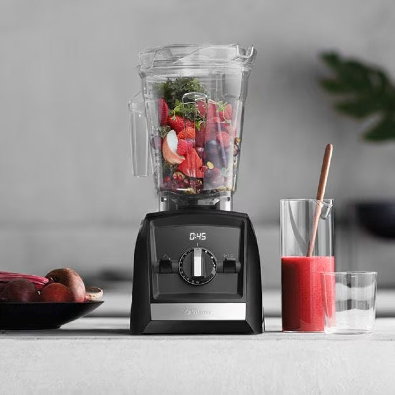 Vitamix Ascent Series A2500i in the kitchen with fruits and vegetables inside and a pitcher of smoothie on the side